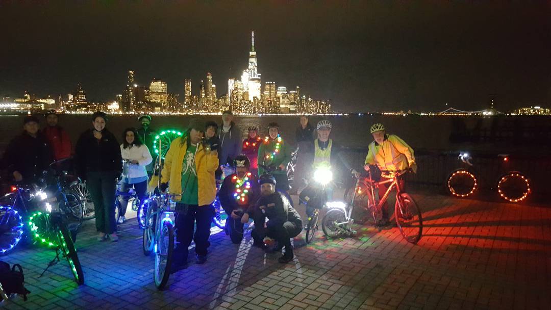 A group of riders stand with their bikes covered in lights in front of the Hudson River and night time New York City skyline.